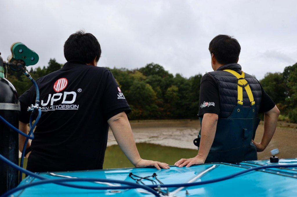 ผู้สวมเสื้อ JPD และเจ้าหน้าที่อยู่ข้างถังน้ำบริเวณบ่อดิน